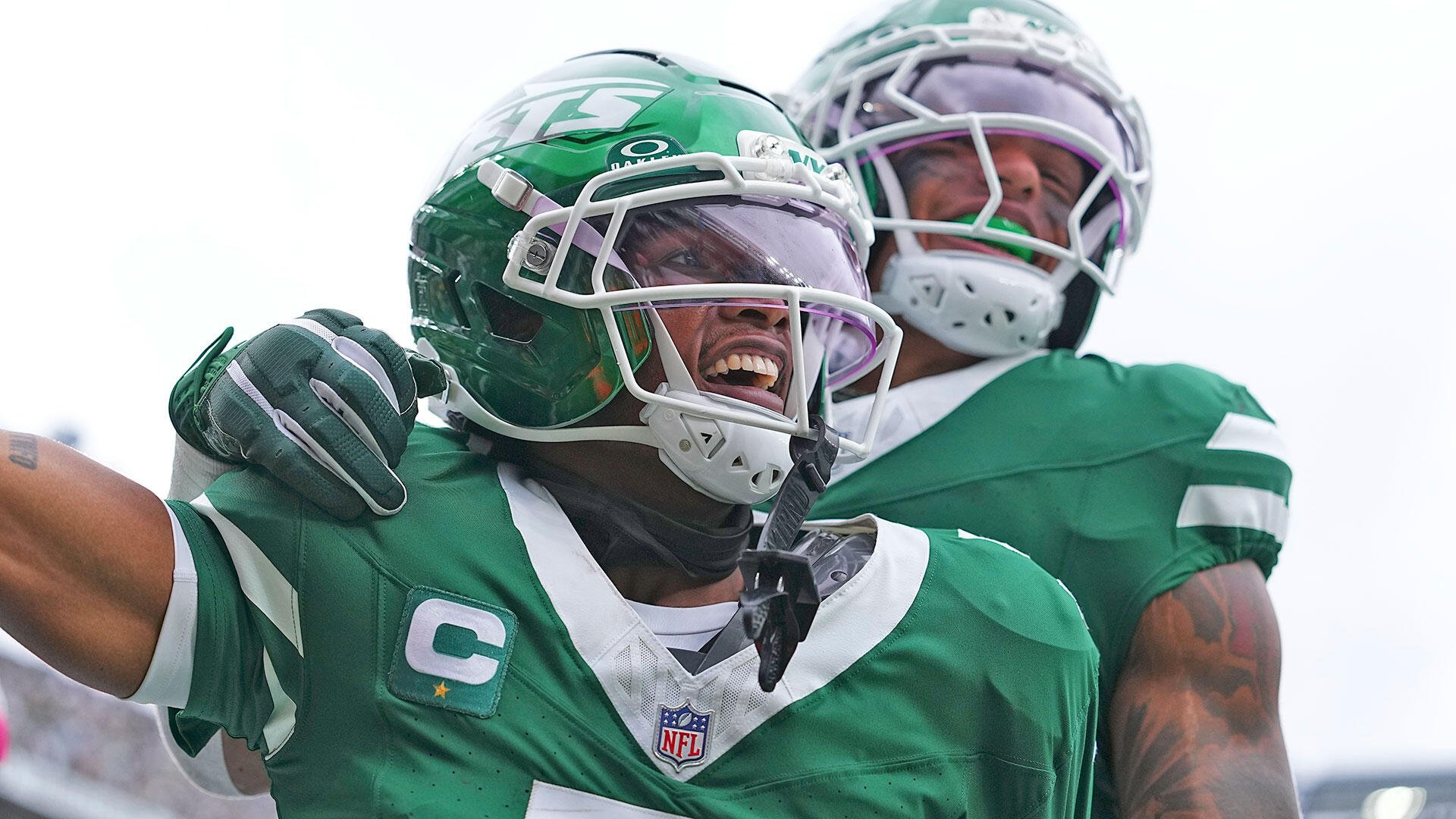 Two football players in green jerseys and helmets celebrate, one gritting his teeth while the other wears a green mouthguard.
