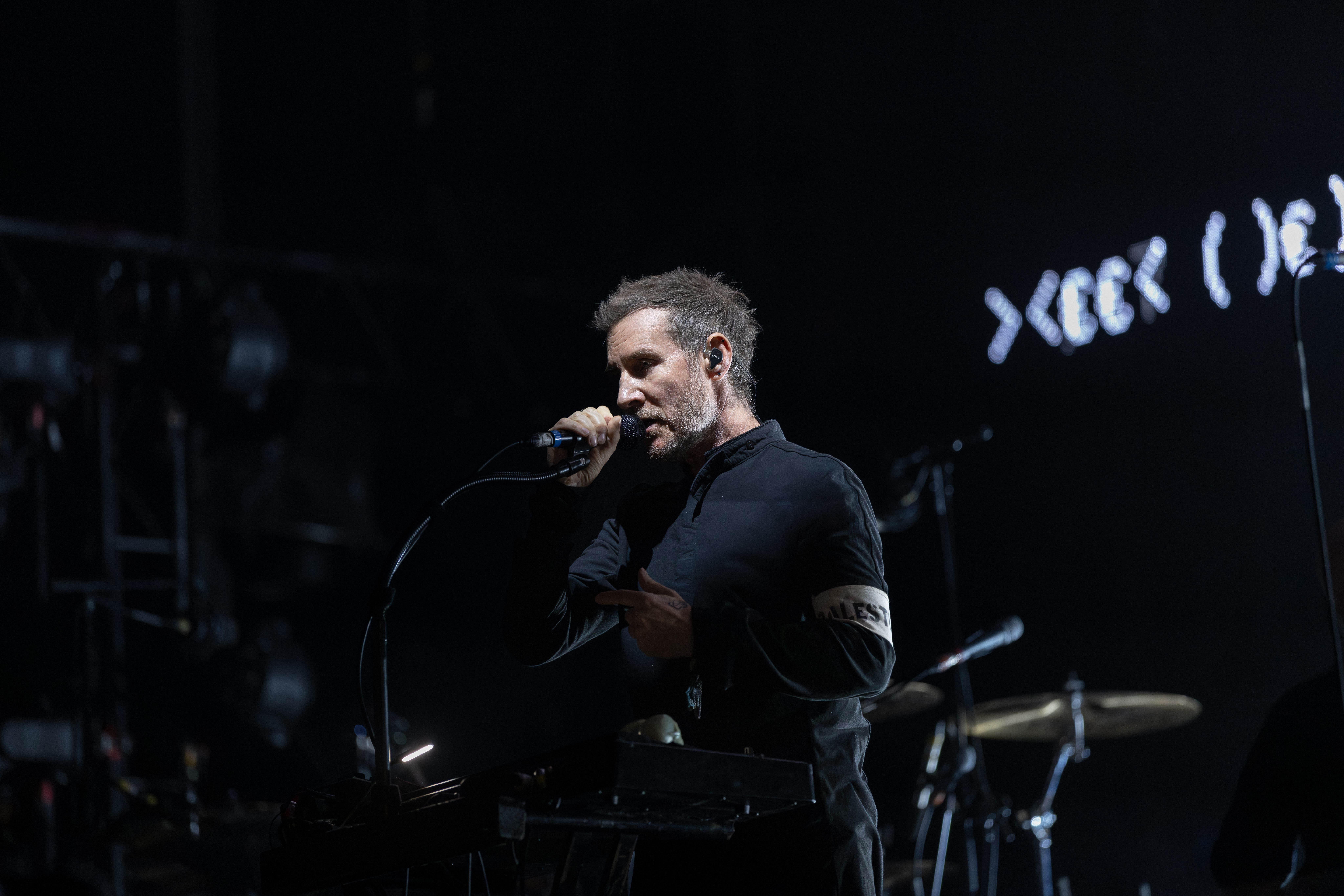 A bearded man in a black shirt sings into a microphone on a dimly lit stage with drum cymbals and stage lights visible in the background.