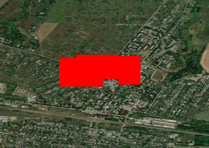 A red rectangular block obscures part of an aerial satellite view showing a town with buildings, roads, green fields, and railway tracks.