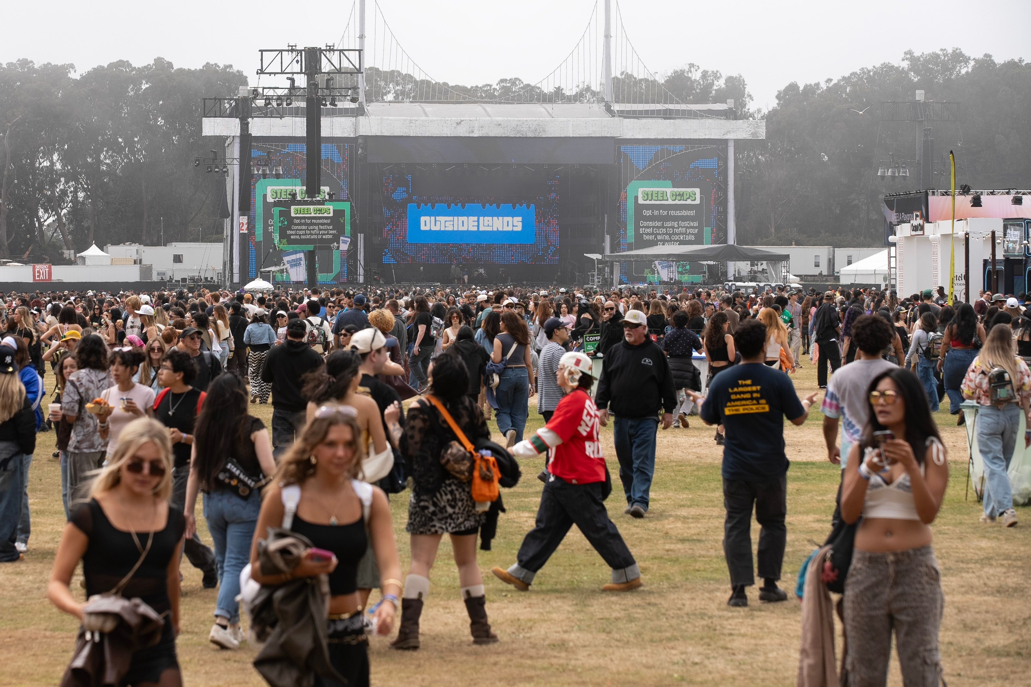 A large crowd gathers on a grassy field in front of an "Outside Lands" festival stage featuring digital screens displaying sustainability messages, with overcast skies and trees in the background.