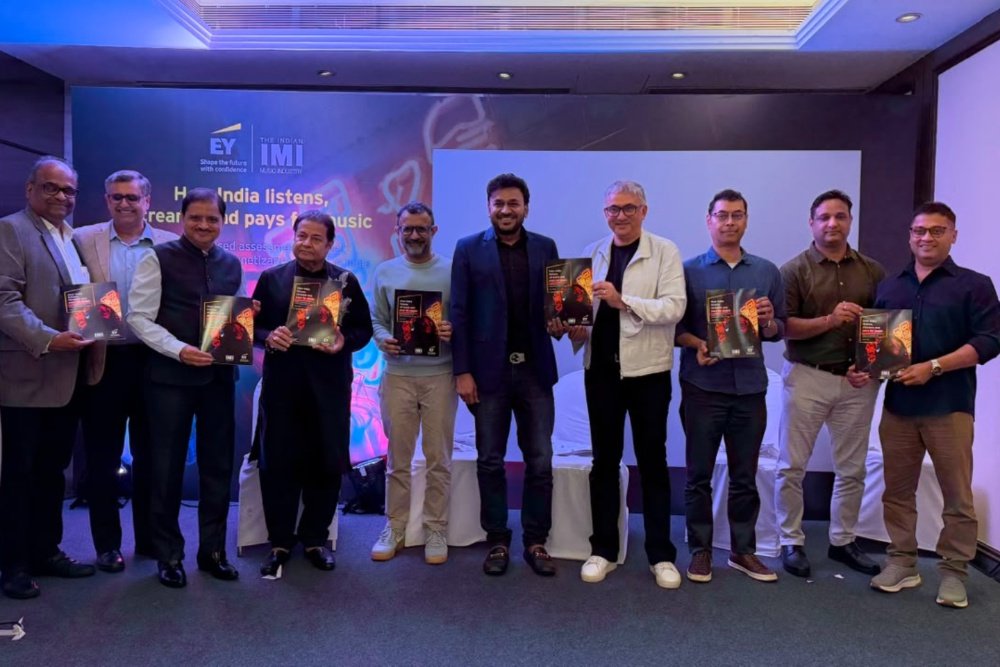Eleven men stand in a line holding copies of an album or book, posing in front of a screen displaying "EY" and "IMI" logos alongside text mentioning India.