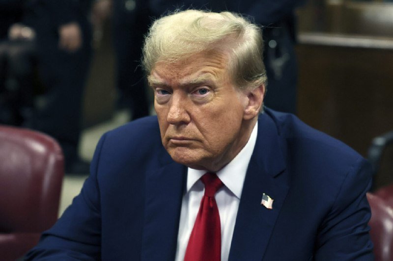 A blonde man in a dark blue suit, red tie, and American flag lapel pin sits with a stern expression at a wooden desk.