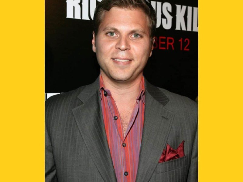 A smiling man in a gray pinstripe suit and red striped shirt stands before a black backdrop with partial white and red text visible behind him.