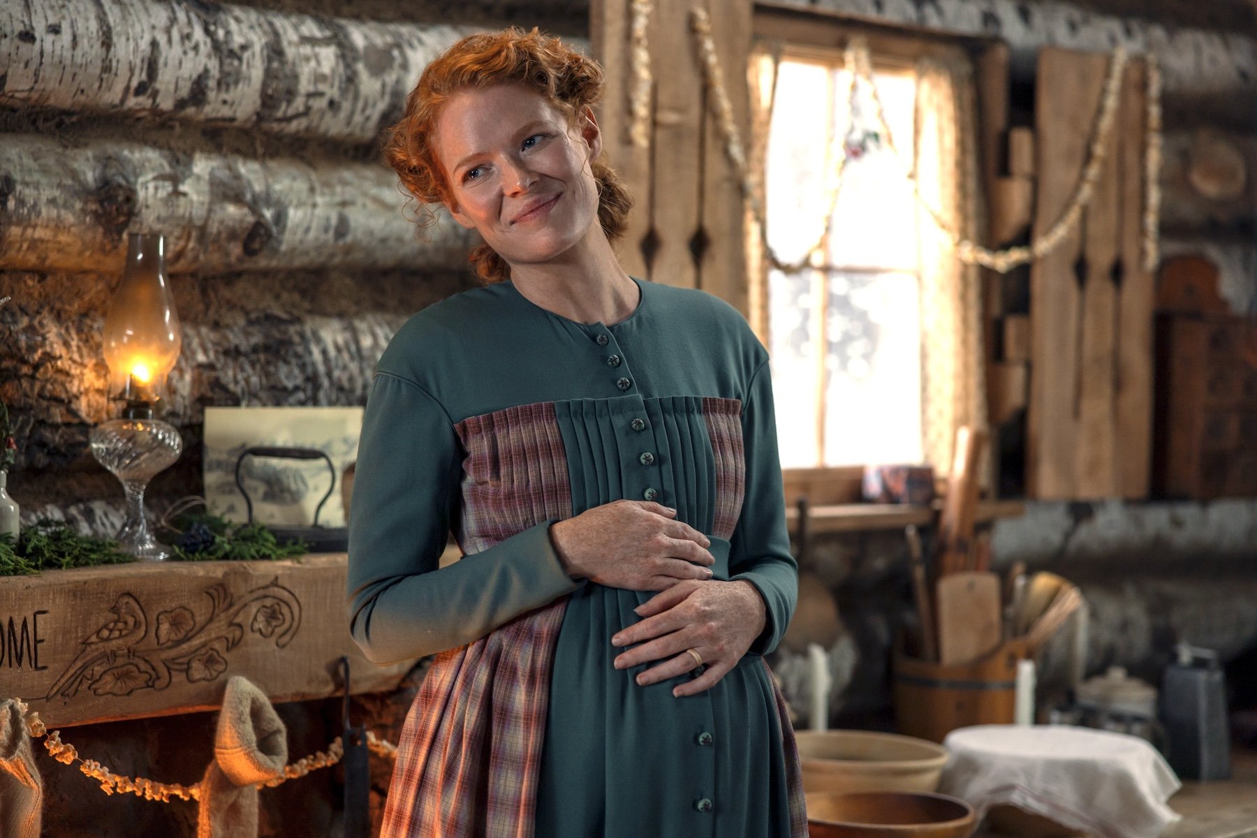 A red-haired woman in a teal and plaid period dress smiles while standing inside a rustic log cabin beside a lit oil lamp.