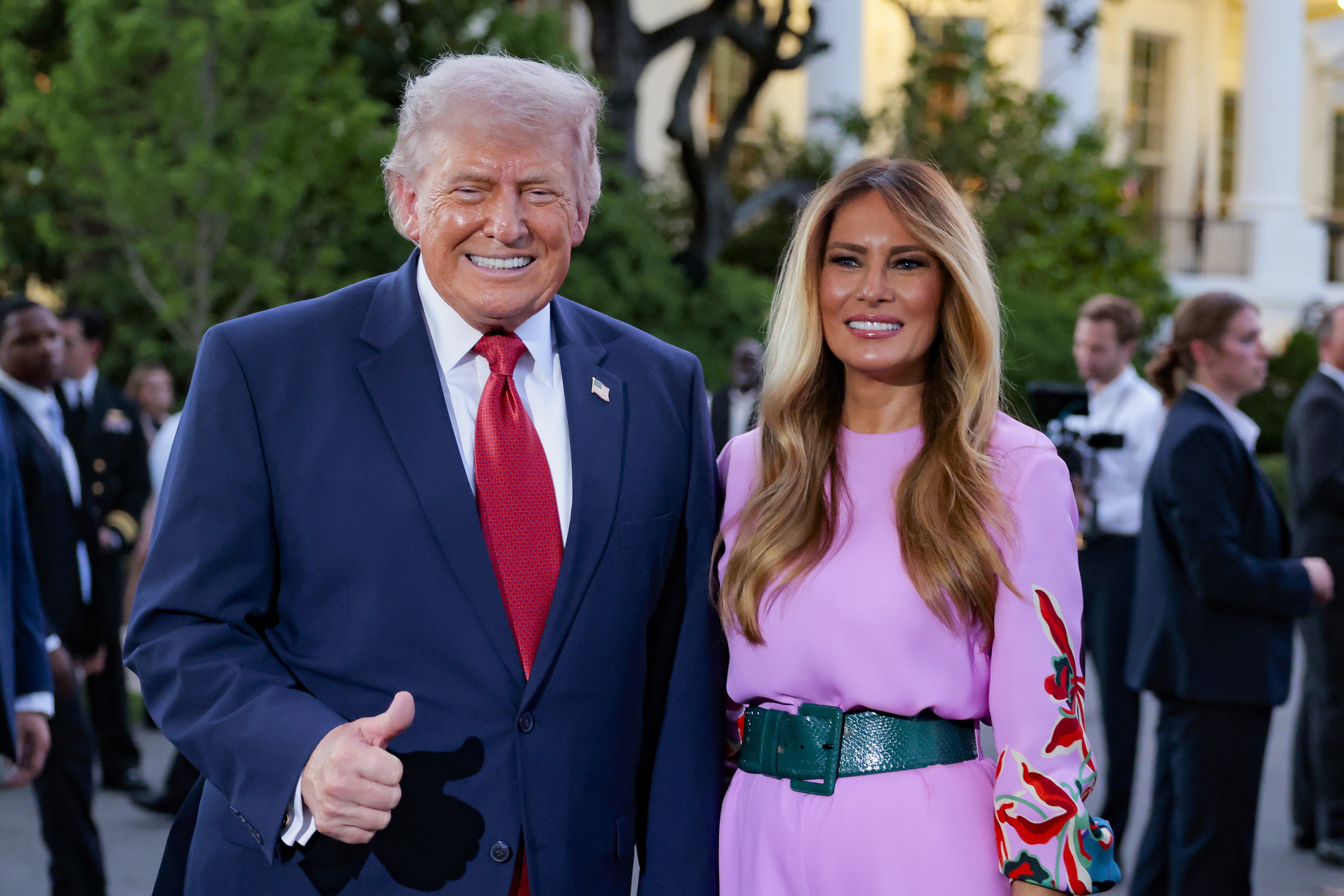 A man in a navy suit with a red tie smiles and gives a thumbs-up next to a woman in a pink dress with a green belt, standing outdoors among other people.