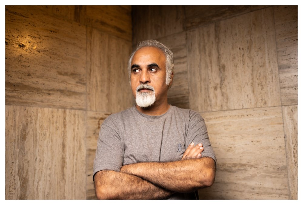 A gray-haired man with a white beard, wearing a gray t-shirt, stands with crossed arms against a beige marble wall, looking off to the side.