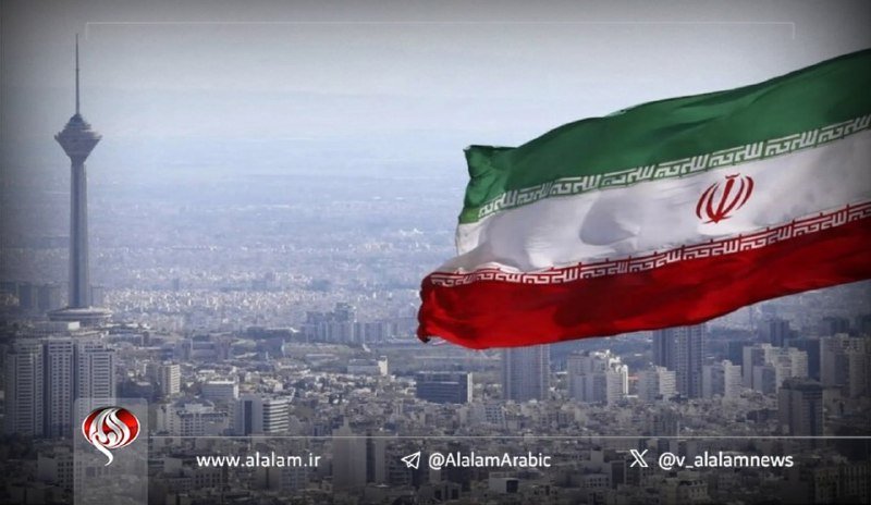 A large Iranian flag waves over a hazy cityscape featuring a tall telecommunications tower.