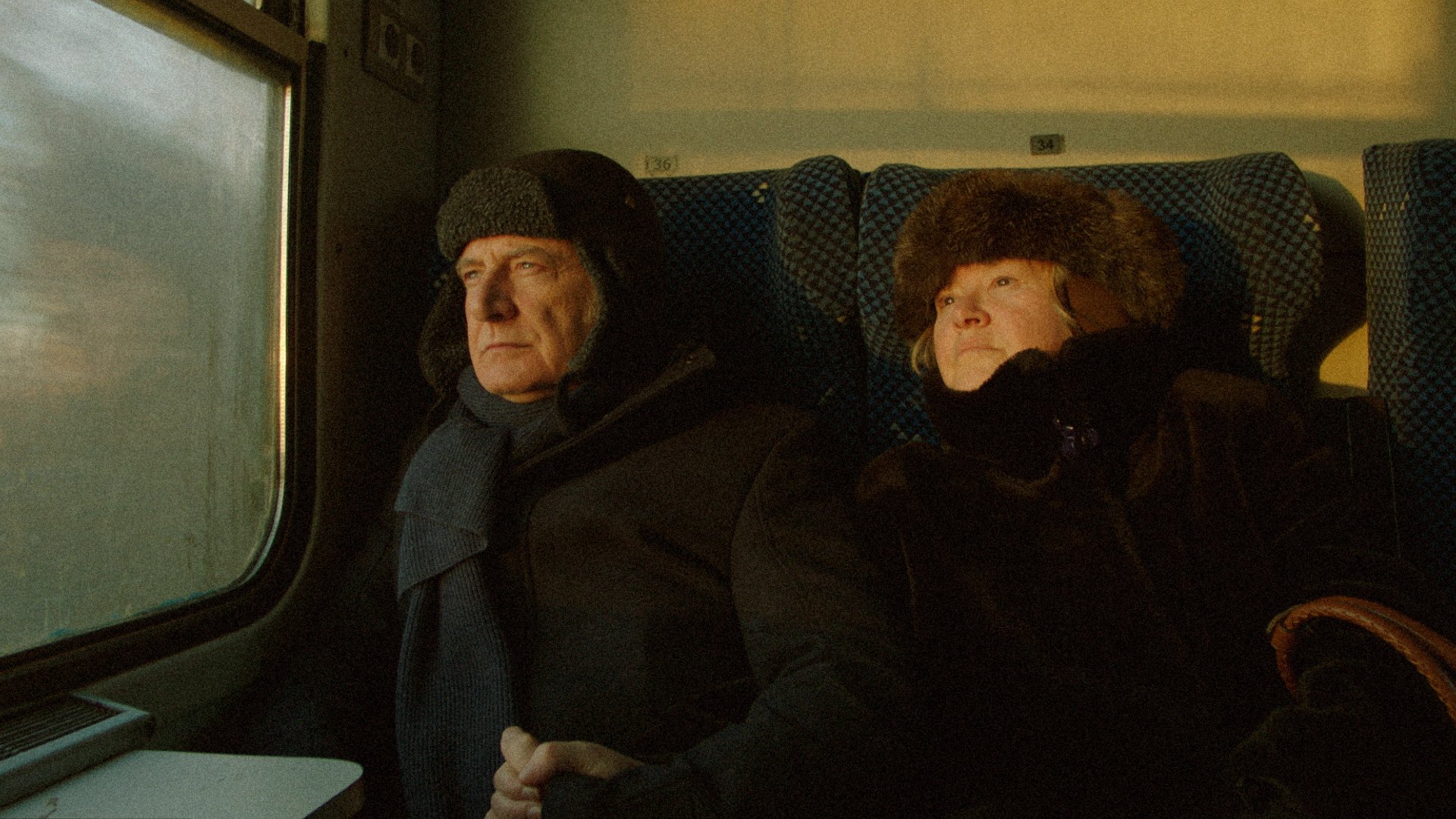 Two older people in winter hats and coats sit beside a foggy train window on patterned seats.