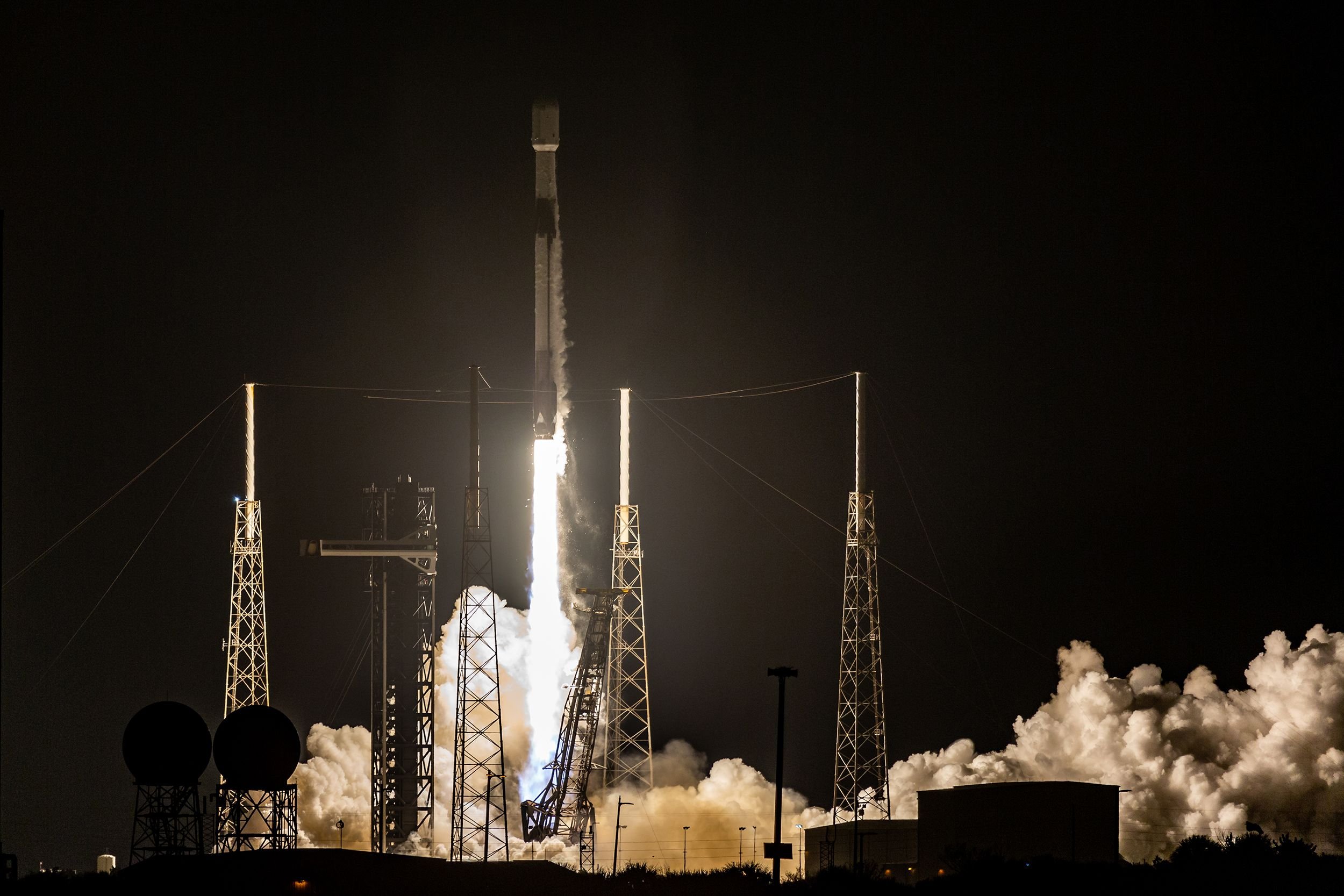 A rocket lifts off at night from a launch pad surrounded by four tall lightning towers, with bright exhaust flames and billowing white smoke illuminating the dark sky.