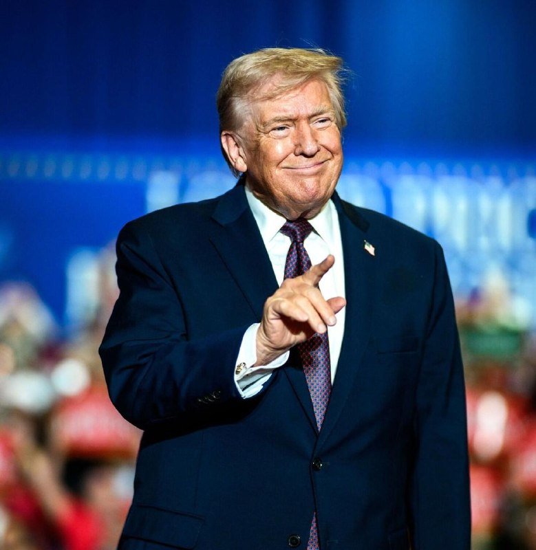 A man with blond hair in a dark suit and purple tie points his finger while smiling on a blue-lit stage before a crowd.