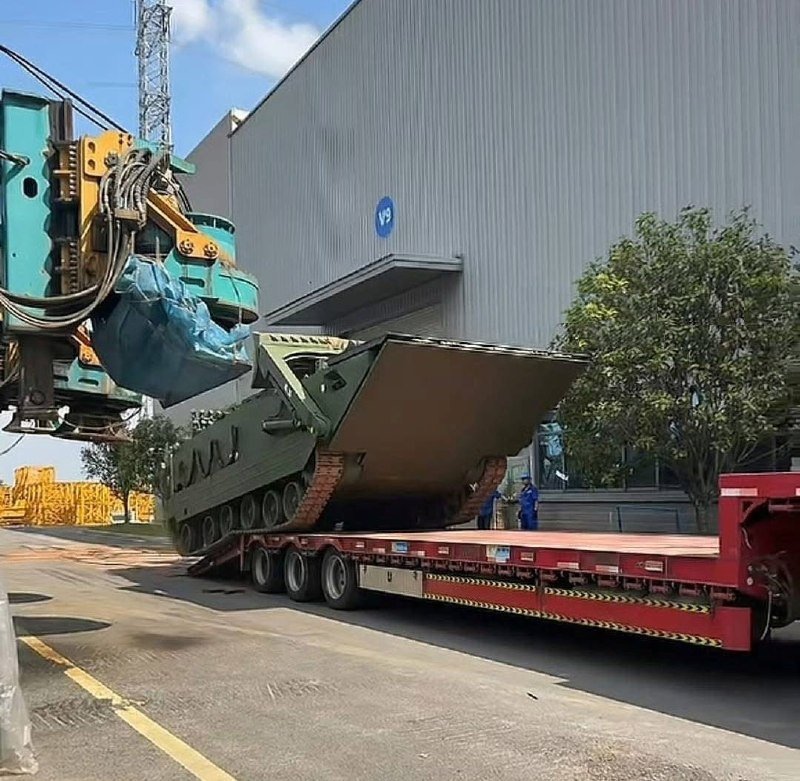 A teal crane lifts a green tracked military vehicle onto a red flatbed trailer outside an industrial warehouse.