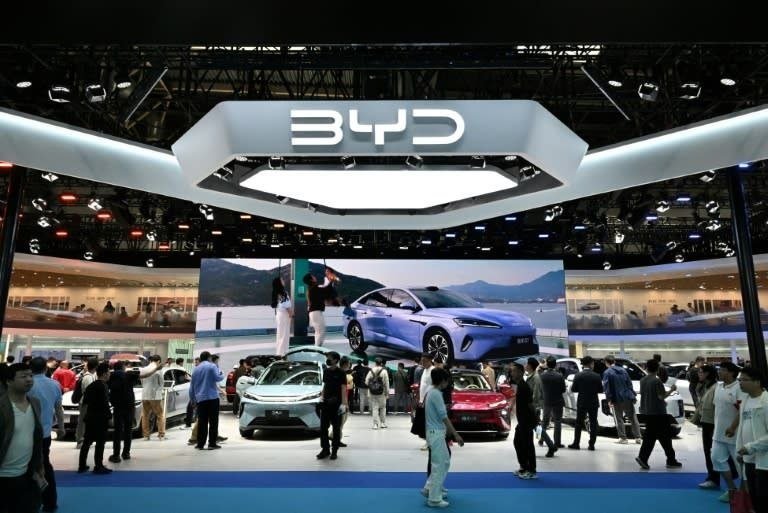 A large "BYD" logo is displayed above an indoor auto show booth where numerous people walk among several displayed vehicles and beneath a large digital screen showing a car.