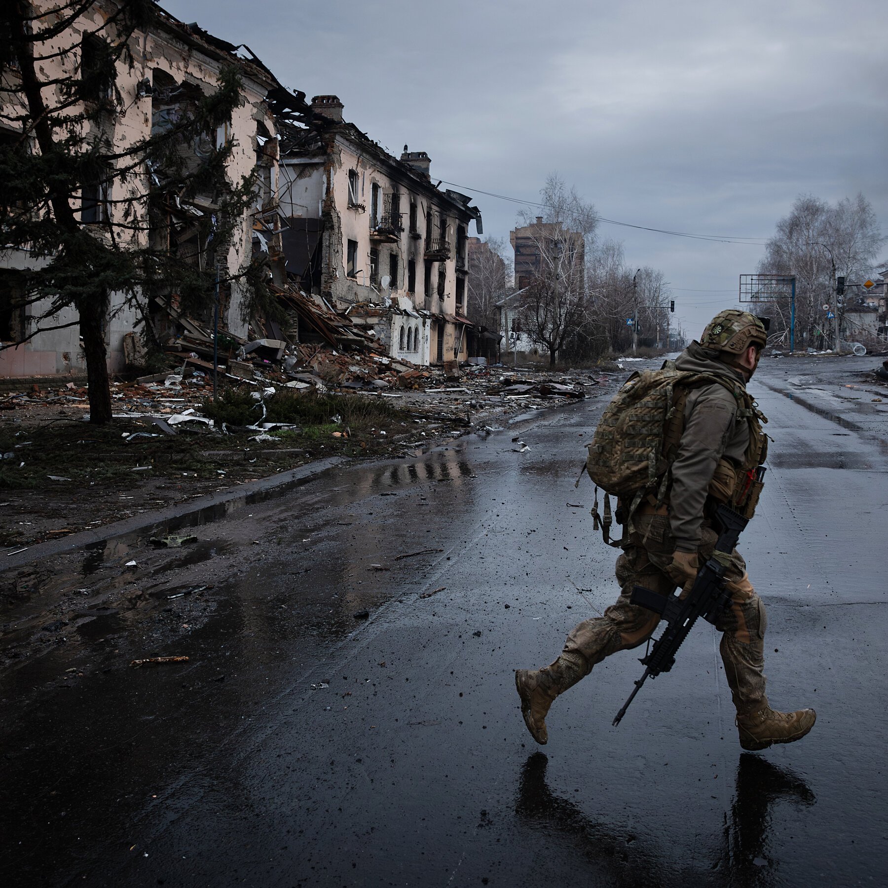 An armed soldier in camouflage runs across a wet street past a heavily damaged multi-story building with debris scattered on the ground.