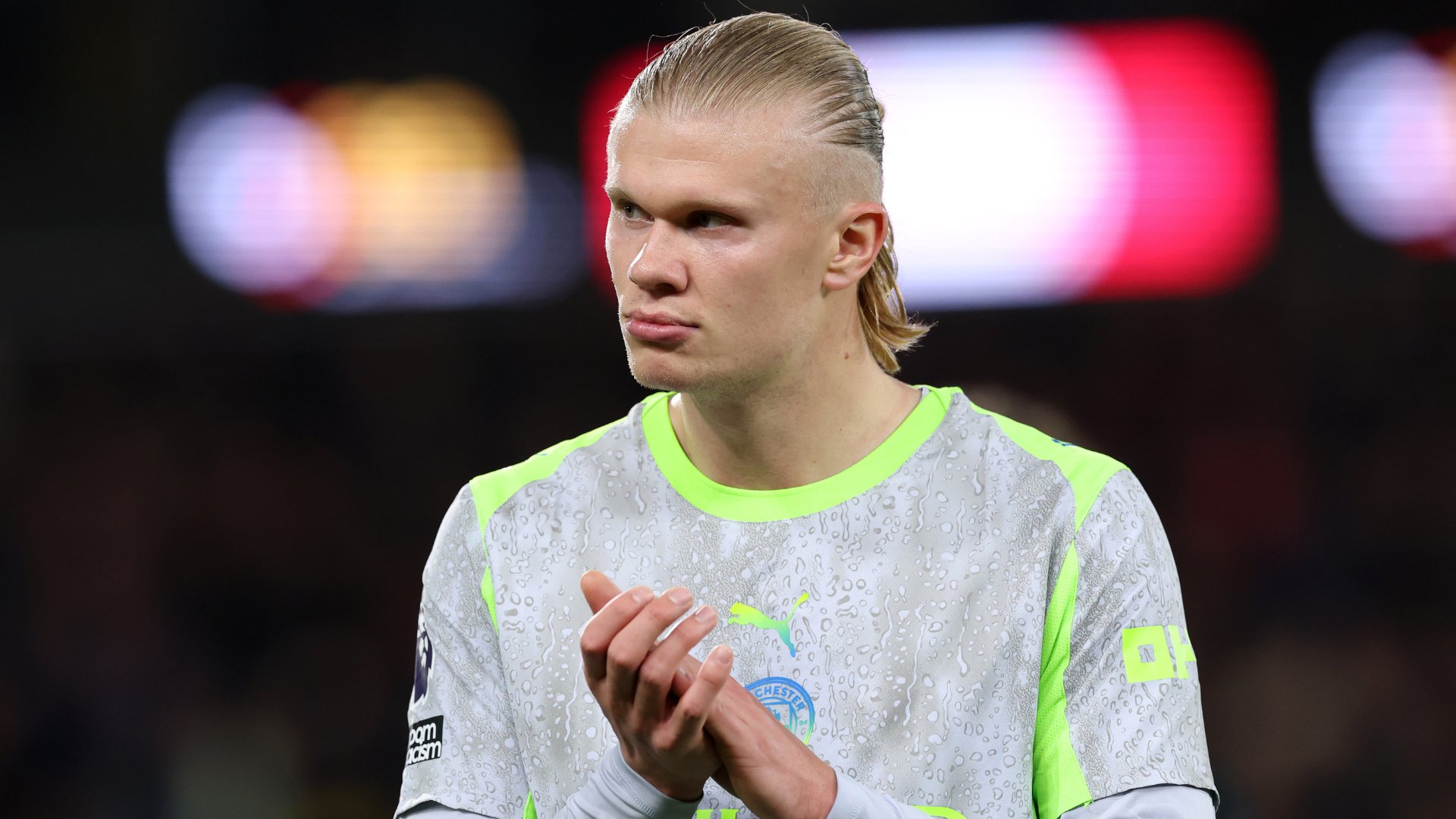 A blonde soccer player in a light gray and neon green Manchester City jersey claps his hands against a blurred stadium backdrop.