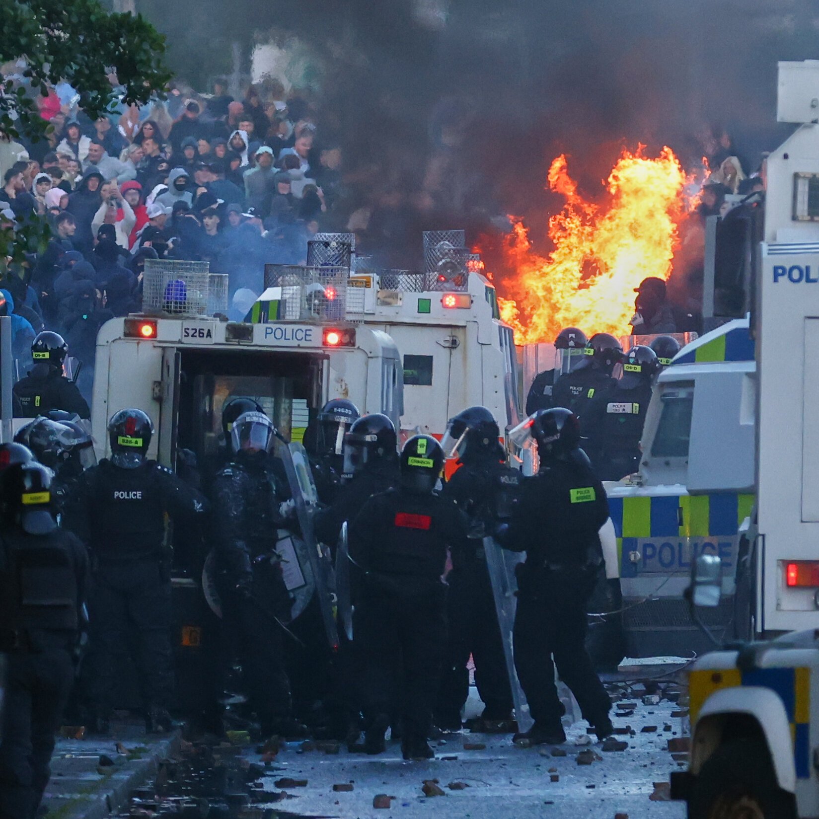 Riot police in helmets and shields stand behind police vehicles as a large fire burns in the street during a confrontation with a crowd.