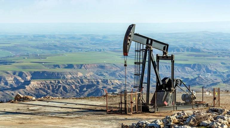 A pumpjack operates on a barren hillside overlooking a vast, eroded landscape of green valleys and badlands stretching to the horizon.