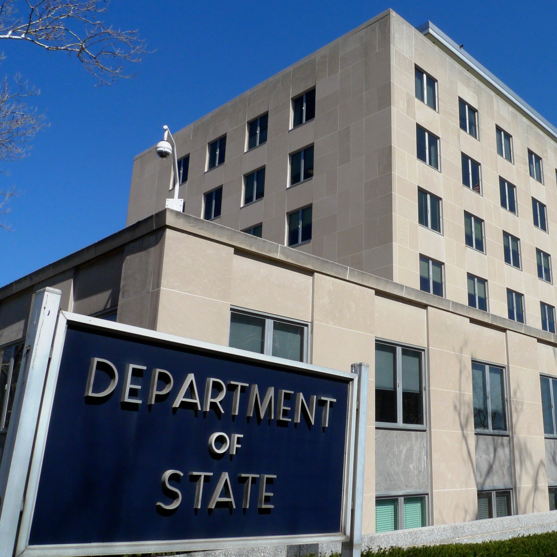 A blue sign reading "Department of State" stands in front of a large, multi-story stone building under a clear blue sky.