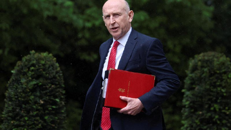 A bald man in a dark suit, white shirt, and red tie carries a red folder while walking past green hedges and foliage.