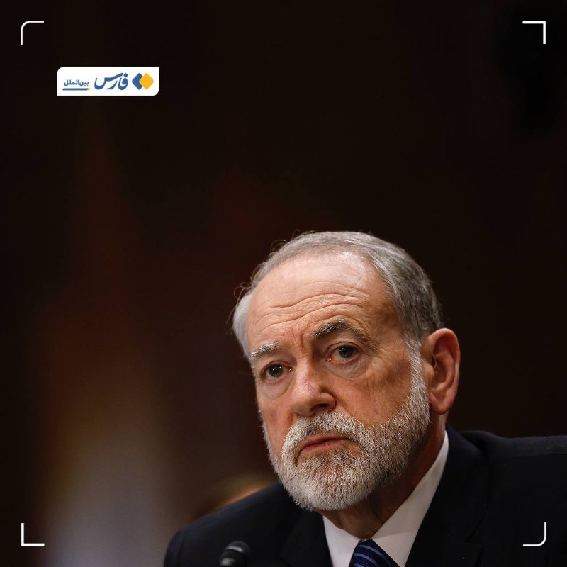 An older man with a gray beard and dark suit sits at a microphone against a dark background, with a Persian-language logo visible in the upper corner.