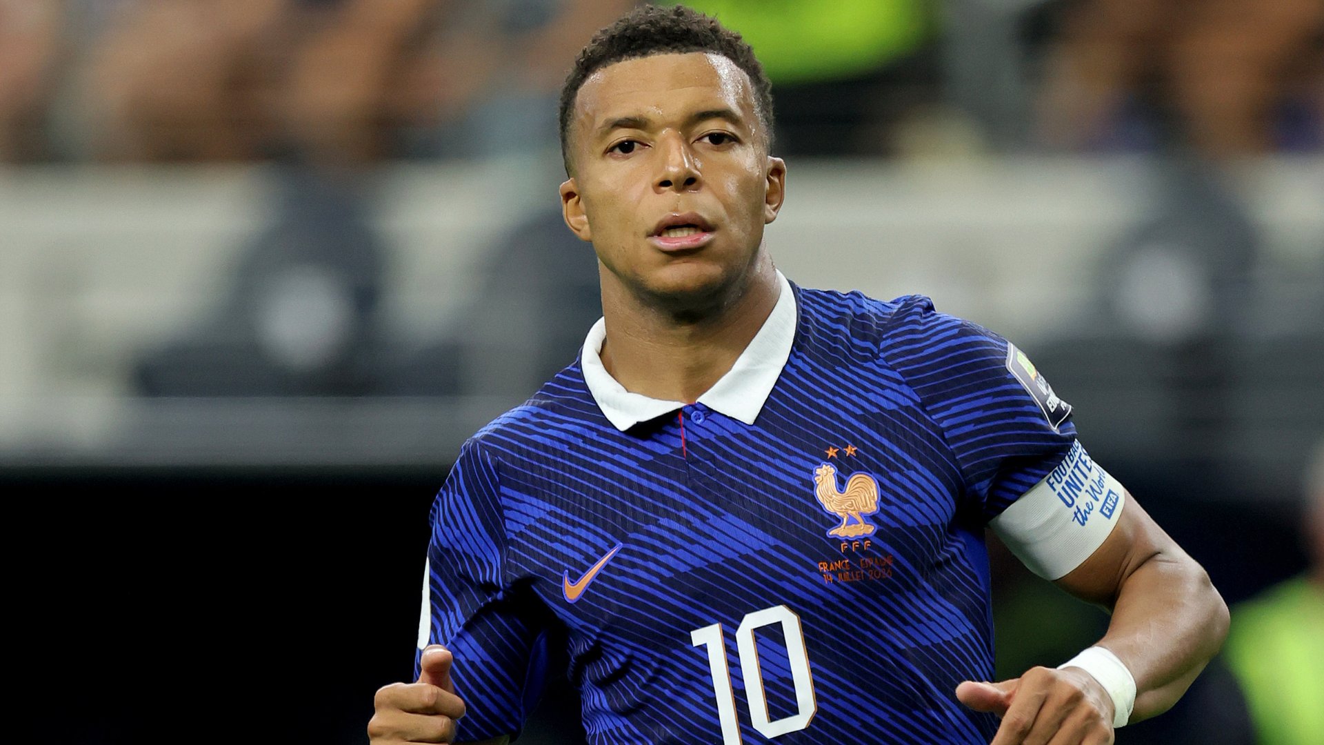 A soccer player wearing a blue France national team jersey with the number 10 runs on the field during a match, a white captain's armband visible on his arm.