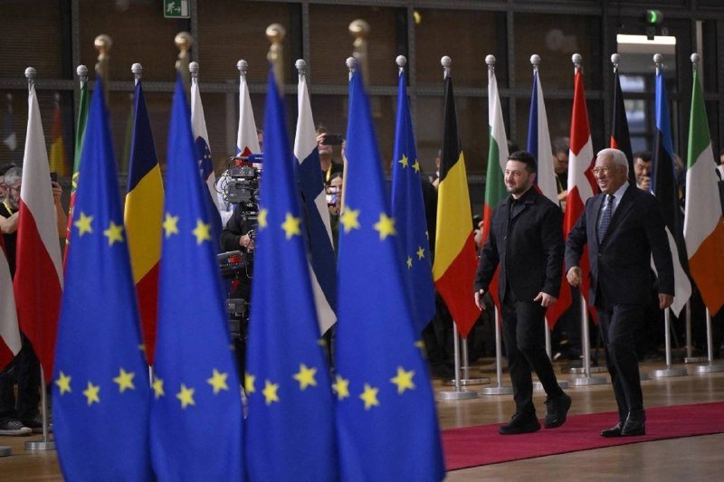Two men in dark suits walk along a red carpet lined with multiple flags, including EU and various national flags, as photographers document the event indoors.