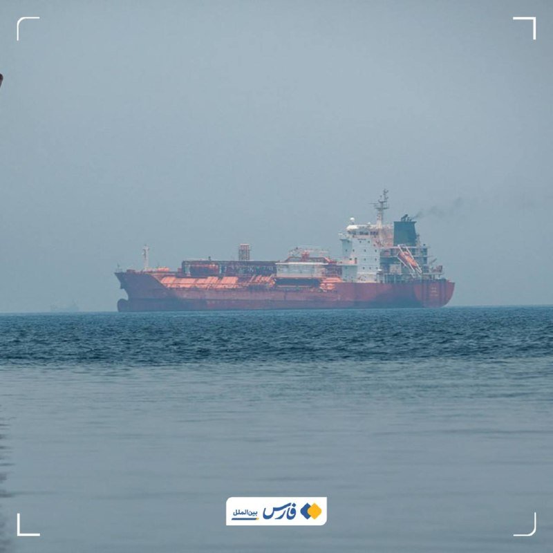 A large red and white tanker ship sails across calm, hazy waters beneath a pale sky.