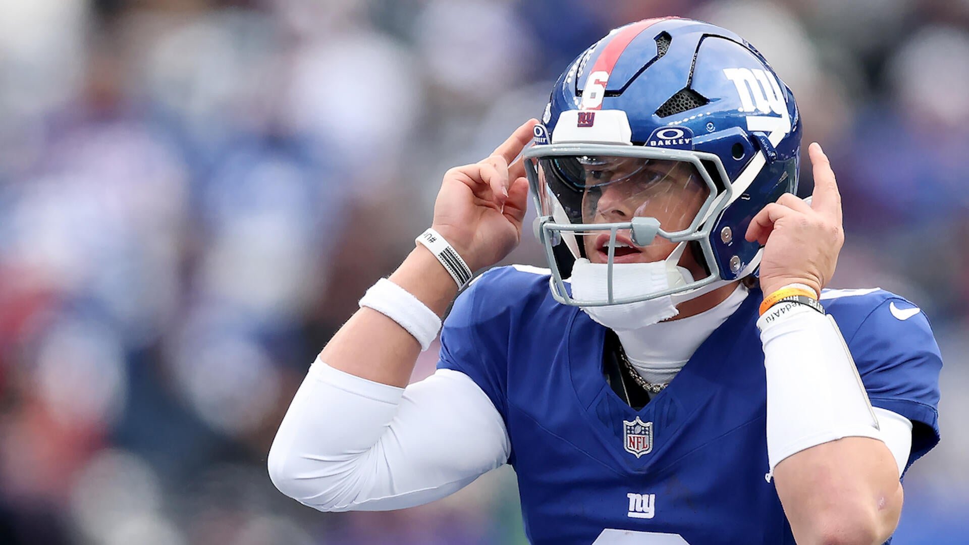 An NFL player in a blue New York Giants uniform and helmet gestures with both index fingers pointing upward on the field.