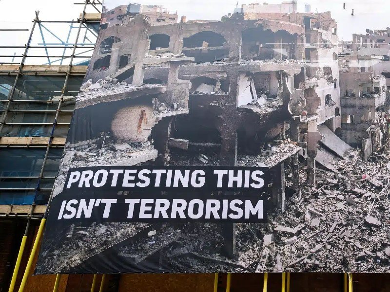 A large banner showing a destroyed multi-story building with the text "PROTESTING THIS ISN'T TERRORISM" hangs in front of a glass building facade.
