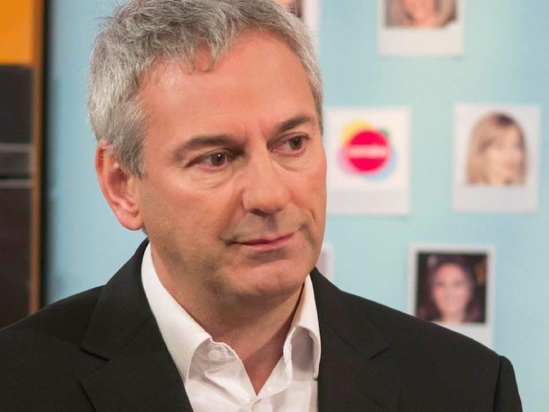 A gray-haired man in a dark suit and white shirt smiles slightly, standing before a light blue wall displaying small portrait photos.