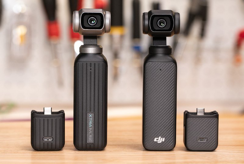 Two handheld gimbal cameras and two modular accessory attachments displayed side-by-side on a wooden surface, with blurred tools visible in the background.