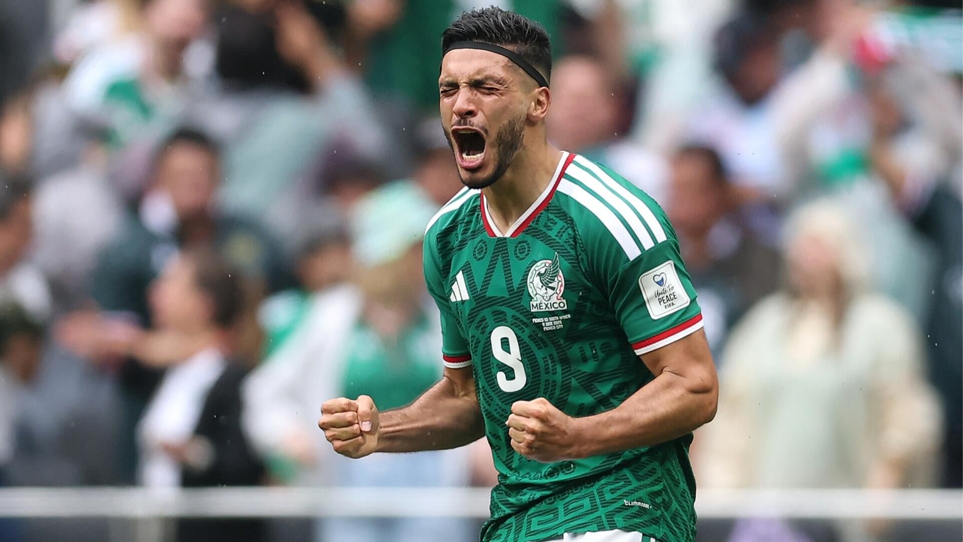 A soccer player wearing a green Mexico jersey with the number 9 clenches his fists and shouts in celebration during a match in front of a crowd.