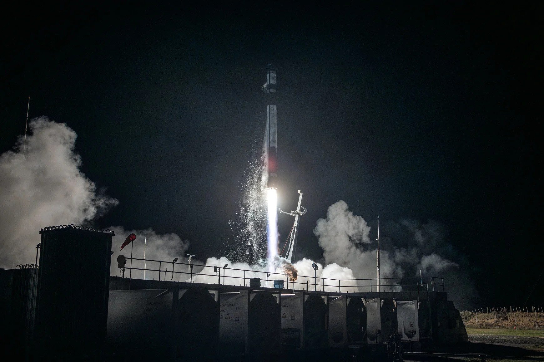 A rocket lifts off from a launch pad at night, its engine flames illuminating billowing smoke and surrounding ground structures.