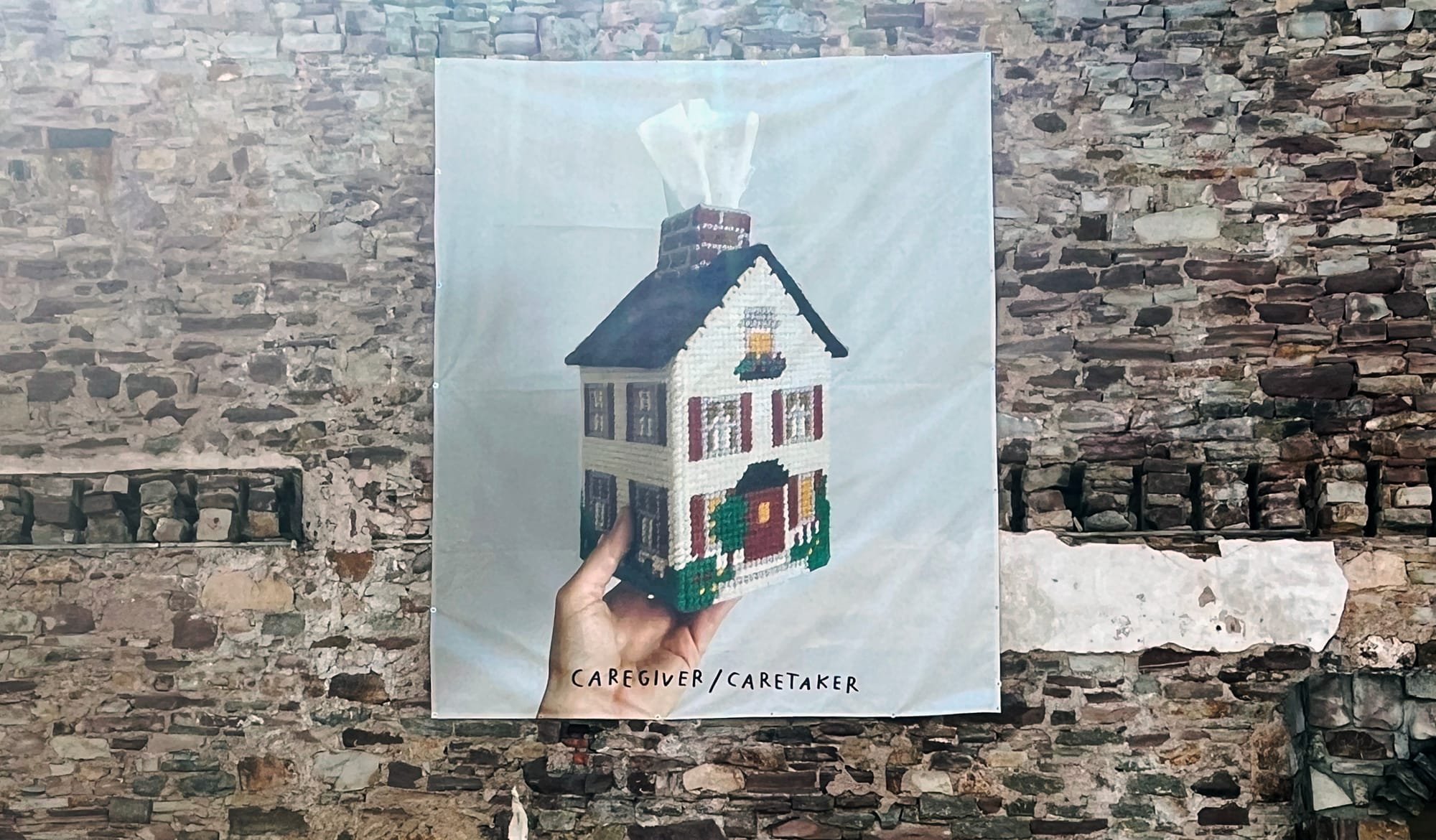 A white banner reading "CAREGIVER/CARETAKER" hangs on a weathered stone wall, displaying a hand holding a tissue-dispensing house.