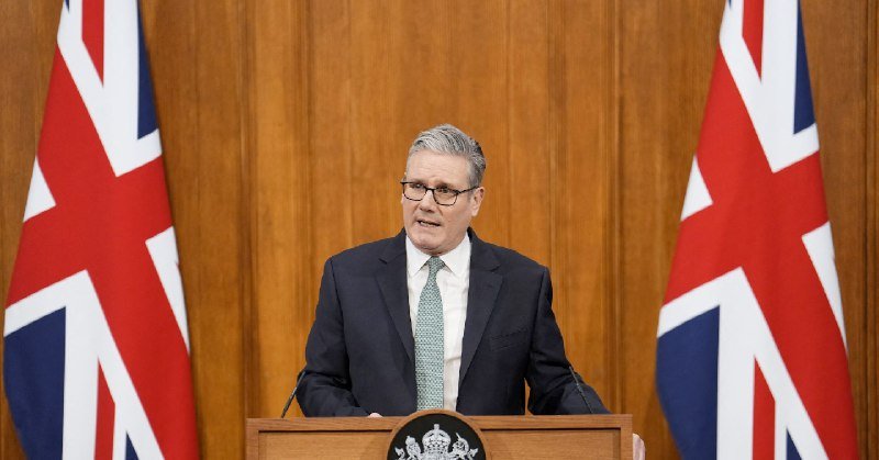 A man in a dark suit and green tie speaks at a wooden podium, flanked by two Union Jack flags.