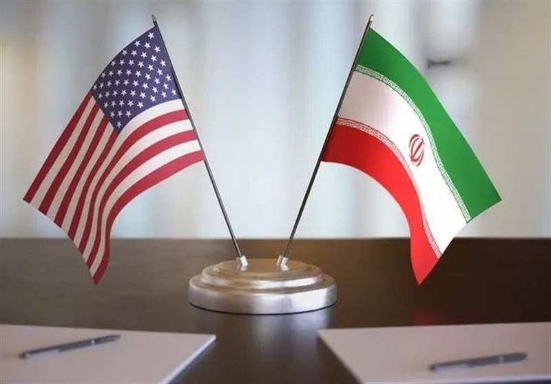 Two small desk flags—the United States and Iran—stand crossed on a shared wooden base atop a dark table, with pens and papers visible nearby.