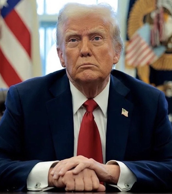 A man with blond hair wearing a dark suit and red tie speaks to reporters holding microphones and recording devices, standing near a wall-mounted screen displaying a presidential seal.