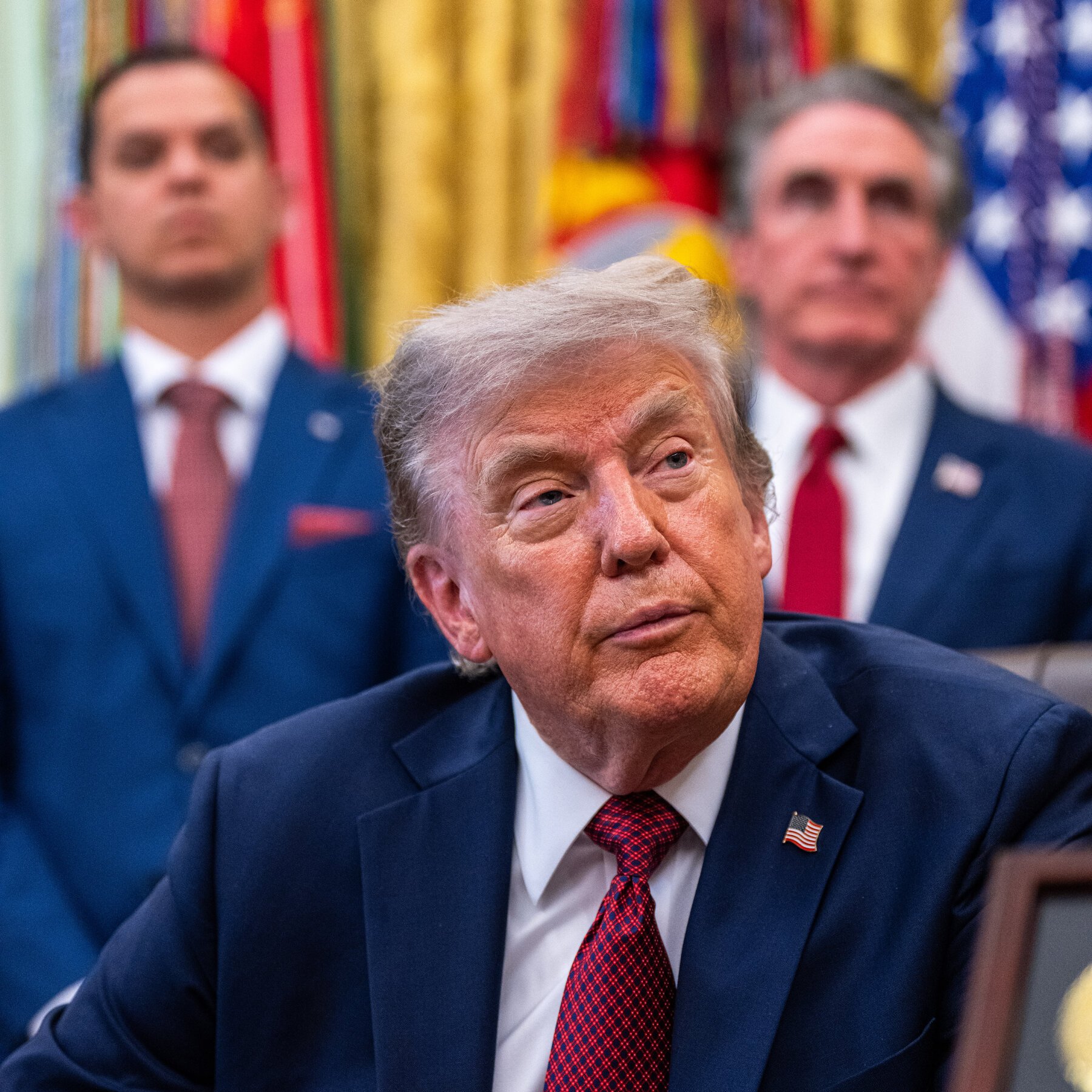 A man with light hair wearing a dark suit, white shirt, and red patterned tie with an American flag pin sits in the foreground, with two suited men and international flags visible behind him.