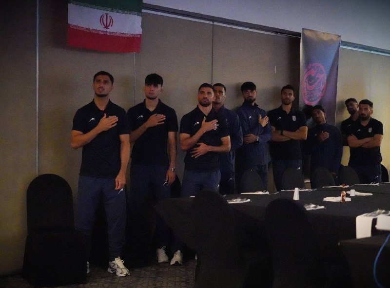 A group of men in dark athletic attire stand solemnly with hands over their hearts beneath an Iranian flag in a conference room setting.