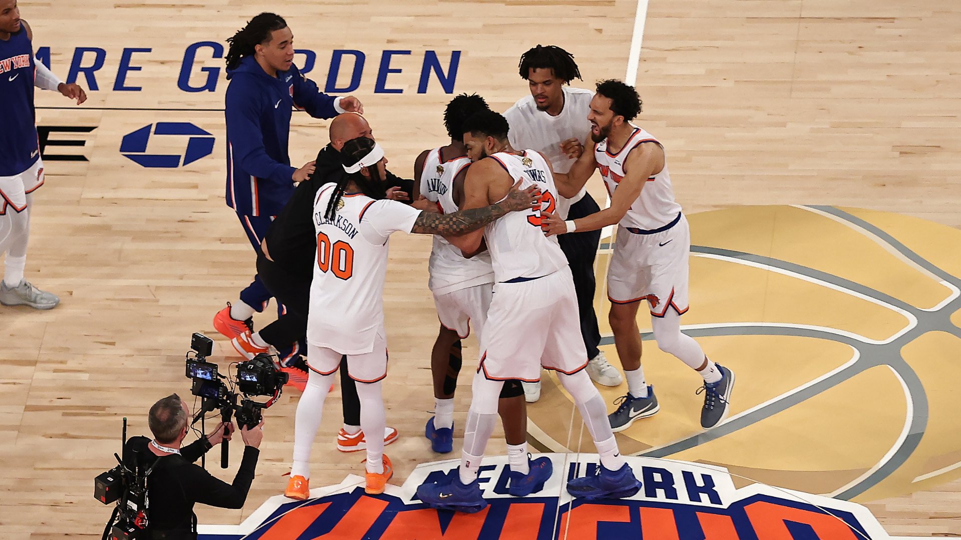 New York Knicks players in white uniforms celebrate together on the basketball court while a cameraman films the scene.