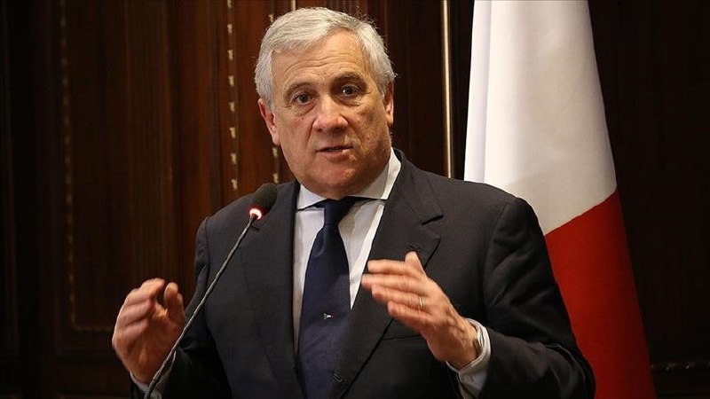 A man with blond hair wearing a navy suit and blue tie speaks indoors, with an American flag and suited individuals visible behind him.