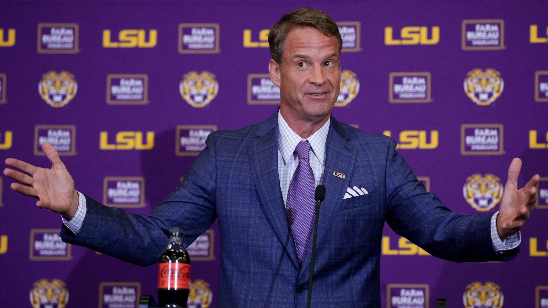 A man in a blue plaid suit gestures with both arms outstretched at a podium in front of a purple backdrop displaying LSU logos and a Coca-Cola bottle.