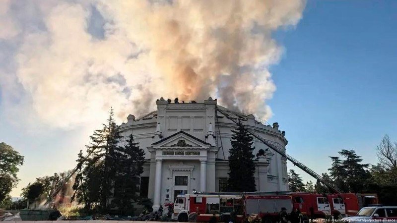 A large white domed building is engulfed in flames, with thick smoke rising into the sky as red fire trucks and firefighters respond at the scene.