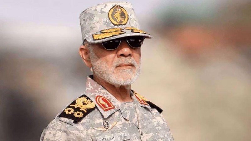 A gray-haired man with a white beard wearing a camouflage military uniform, cap with an emblem, and dark sunglasses stands outdoors.