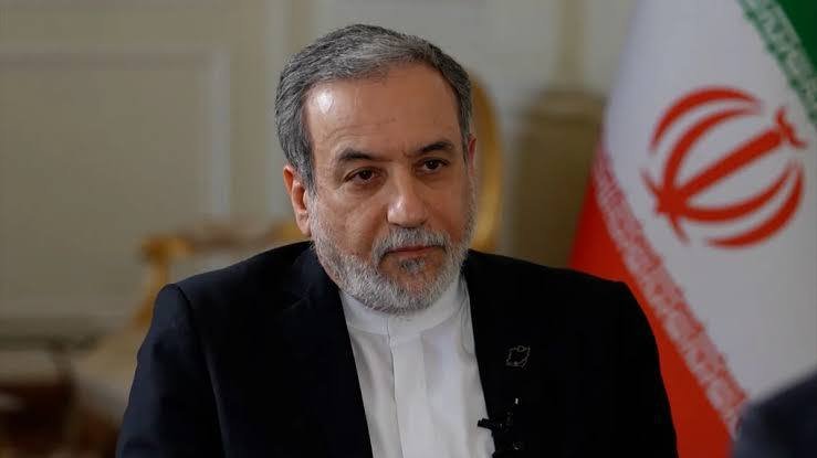 A gray-bearded man in a dark suit and white shirt sits beside an Iranian flag during what appears to be a televised interview setting.