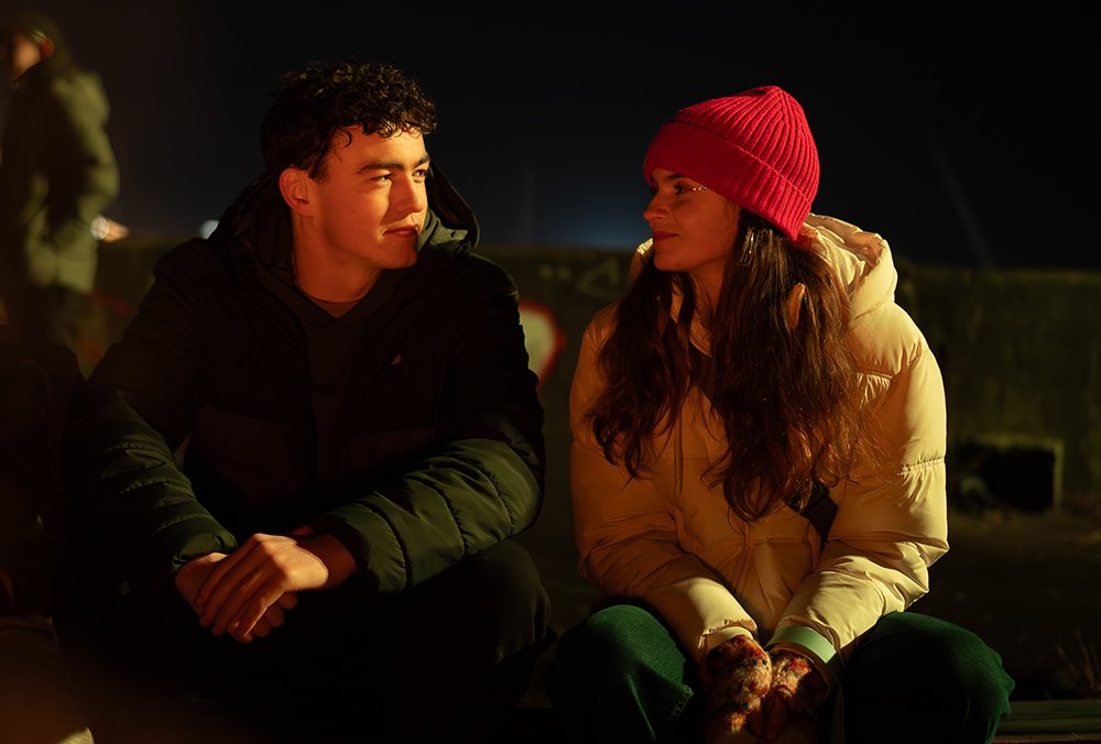 A young man and woman sit side by side outdoors at night, gazing at each other in warm firelight; he wears a dark puffer jacket, she wears a cream coat and red beanie.