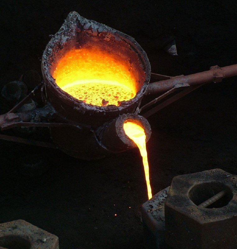 Molten metal pours from a spout of a heated crucible into a mold below, glowing bright orange against a dark industrial background.