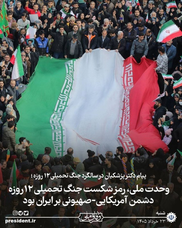 An Iranian flag is held above a large crowd of people, many of whom are waving smaller flags and holding up phones, as Persian text appears on screen.