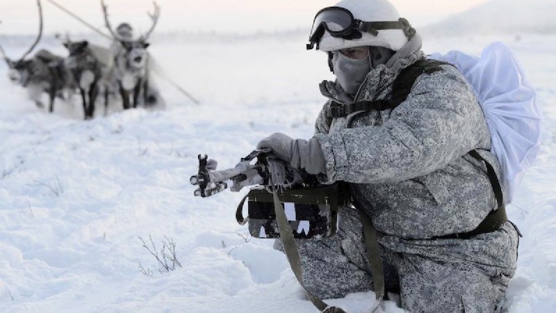 A camouflaged soldier kneels in snow holding a rifle, while a figure drives a reindeer-drawn sled in the background across a snowy, treeless landscape.