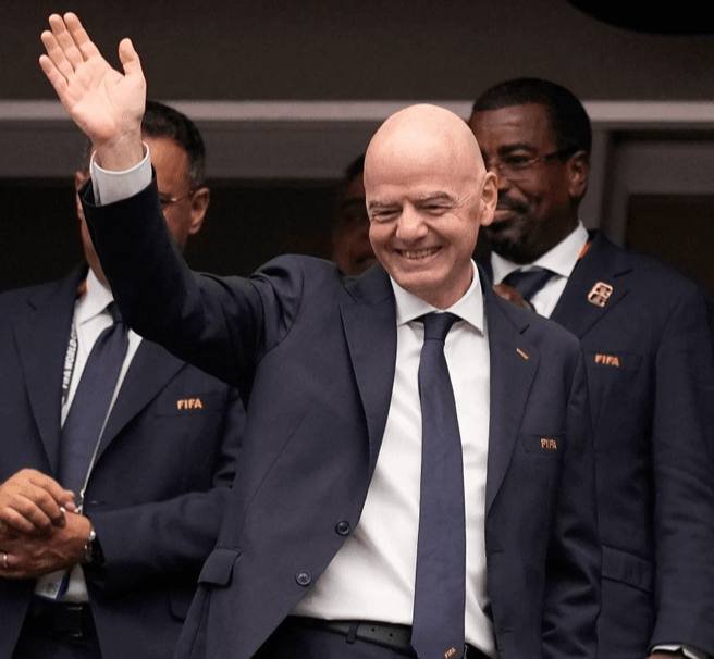A smiling bald man in a dark suit waves his right hand while standing beside other men wearing FIFA-branded suits.