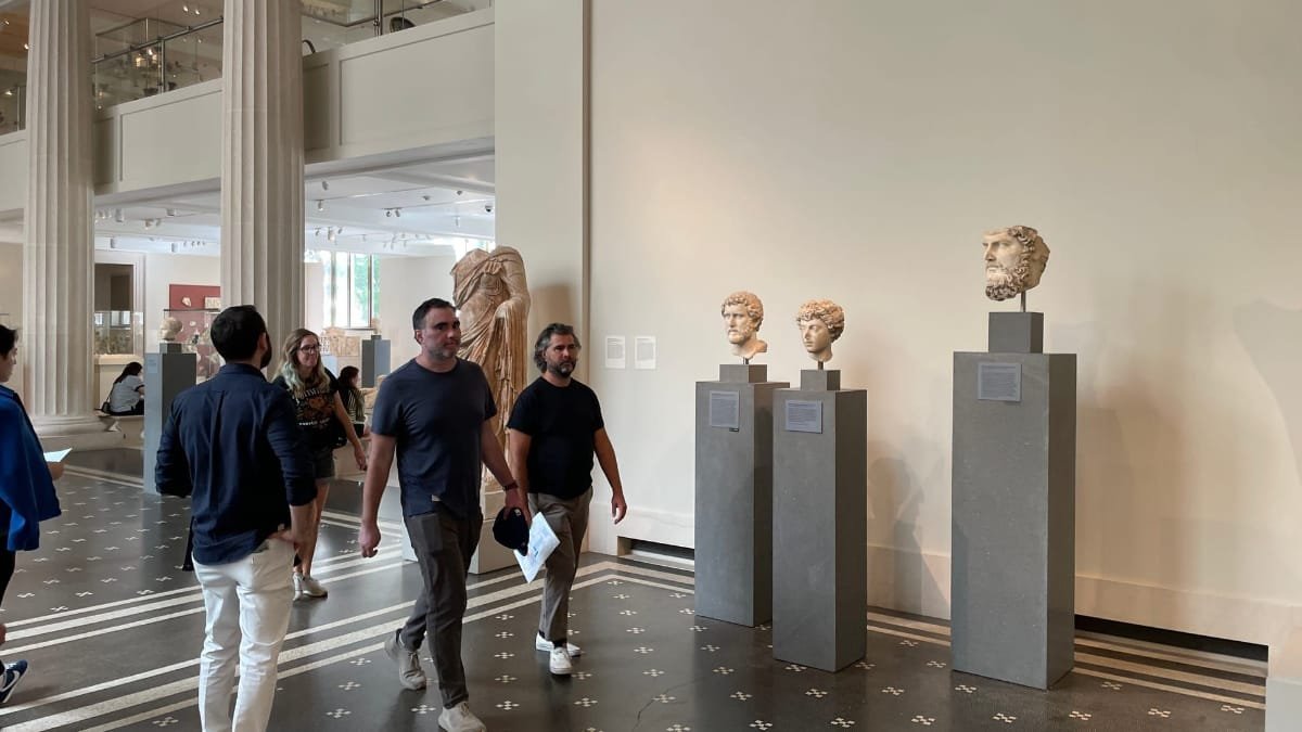 Visitors walk through a museum gallery past ancient stone busts and a draped figure displayed on gray pedestals.